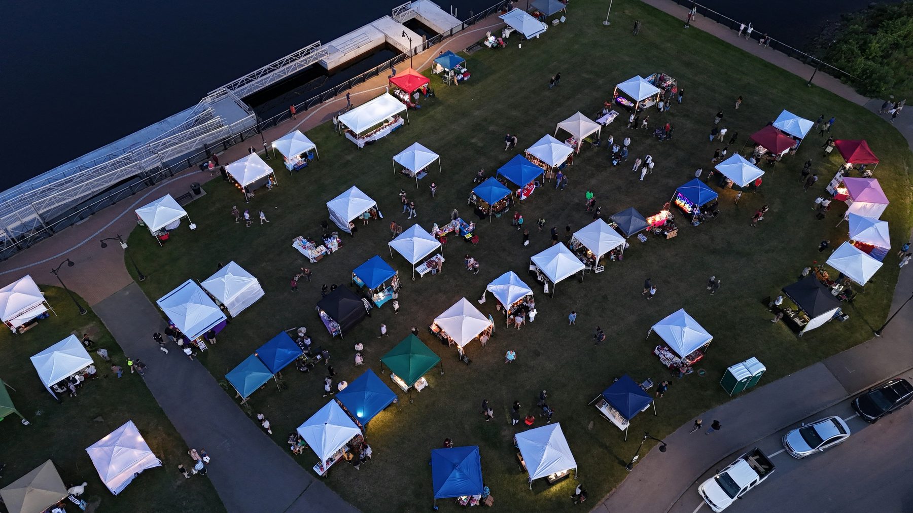 Outdoor pop-up event with many tents, people, and nearby parked cars
